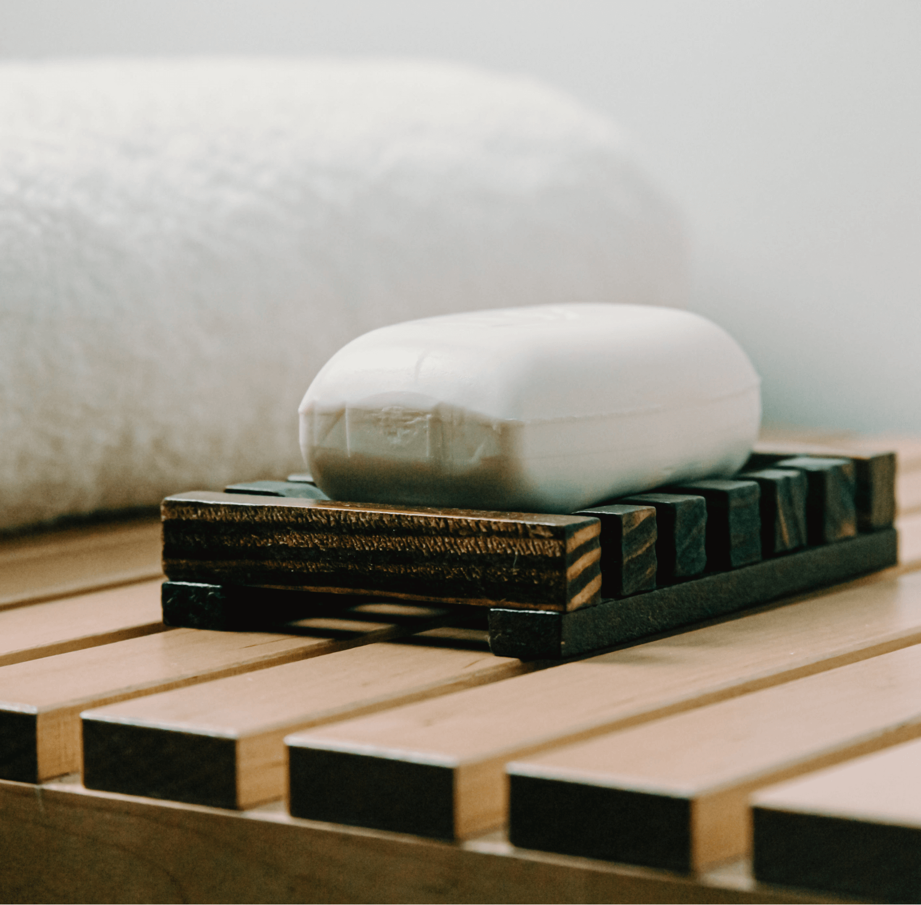 A white bar of soap resting on a dark wooden slatted soap dish, placed on a light wood slatted surface with a soft white towel blurred in the background.
